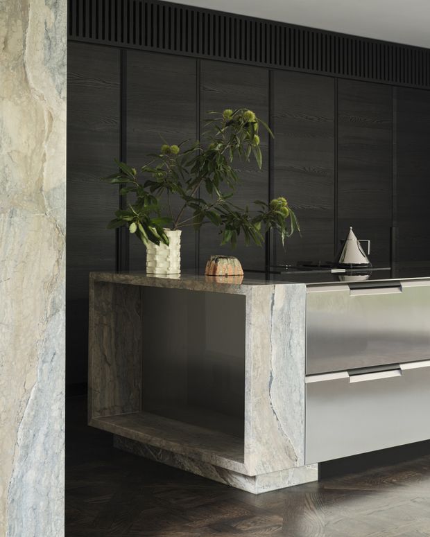 A refined contemporary kitchen featuring a substantial stone-clad island with an open display niche, dark timber wall cabinetry, and sleek grey drawer fronts. A black integrated ventilation grille runs above the cabinetry, while potted greenery and sculptural objects add a natural decorative touch. The space has dark timber flooring and a sophisticated, moody palette.