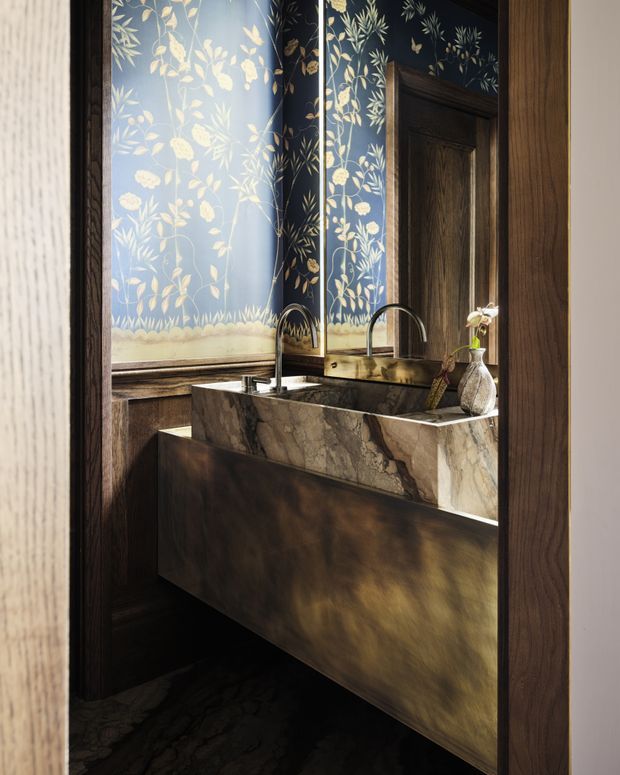 A dramatic, richly detailed bathroom vanity area viewed through a timber-framed opening. The room features a dark floating vanity with a thick veined natural-stone basin and benchtop, dual arched metal taps, dark timber wall panelling and door, and a botanical floral wallpaper panel in deep blue with pale foliage. Warm directional light highlights the stone and brushed metallic vanity front.