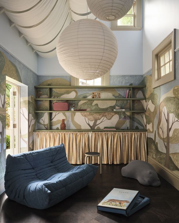 A richly styled compact lounge or reading room with a sculptural deep-blue upholstered lounge chair, dark timber flooring, and a dramatic floor-to-ceiling woodland mural in muted green, taupe, cream, and brown tones. Open dark shelving spans the rear wall above a pleated fabric-fronted built-in cabinet. Large spherical paper pendant lights and layered draped ceiling fabric create a playful, theatrical atmosphere, while timber-framed windows and a glazed door bring in natural light.