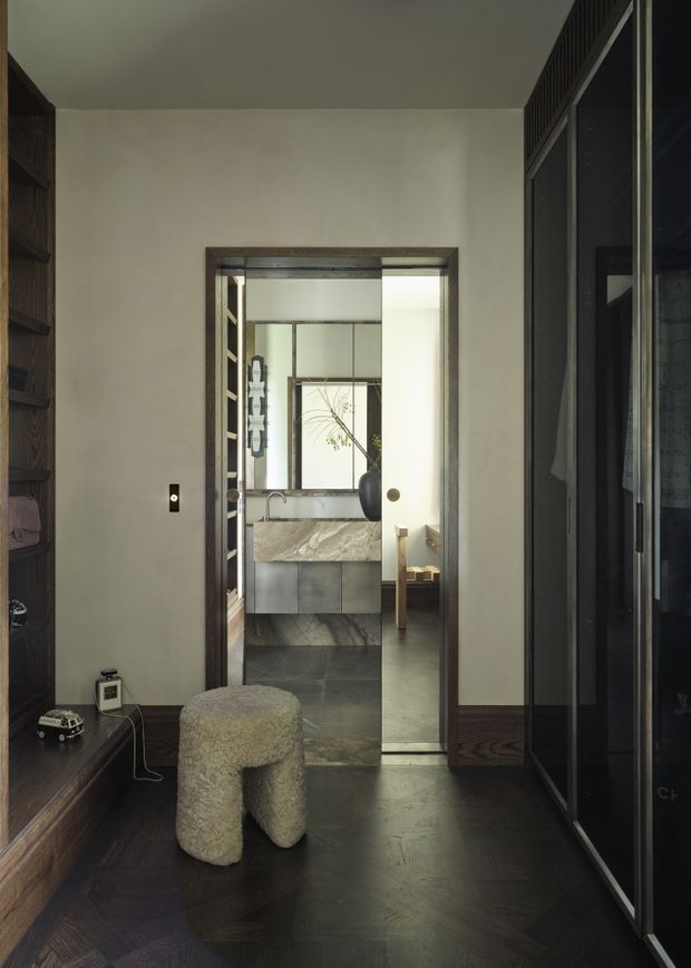 A moody, refined interior view looking through a dark timber-trimmed doorway toward a compact bathroom or vanity area. The foreground has dark timber flooring, built-in dark shelving and cabinetry, a textured boucle stool, and full-height dark-framed glazing or wardrobe doors. Beyond the doorway is a stone vanity with integrated basin, mirrored surfaces, grey cabinetry, and a freestanding decorative branch. The space is softly lit with a restrained neutral palette and strong material contrast.
