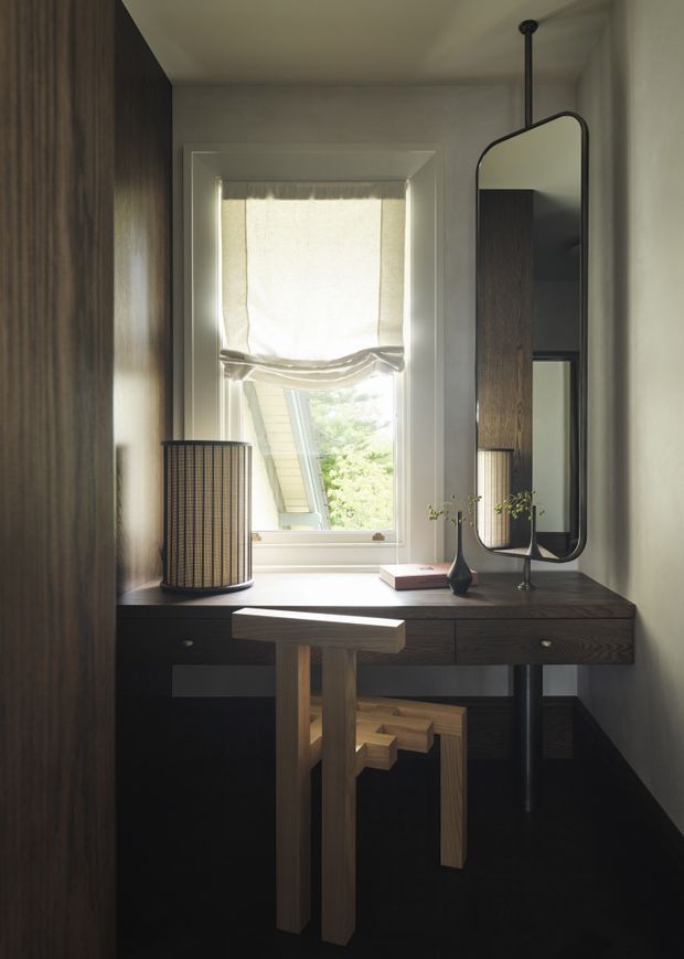 A compact, refined home office or dressing workspace centered on a dark timber built-in desk beneath a tall window. The space includes a slim suspended-framed mirror, sculptural timber stool, dark wood cabinetry, a woven cylindrical table lamp, and minimal decorative objects. Soft daylight filters through a Roman shade, creating a warm, intimate and sophisticated atmosphere.