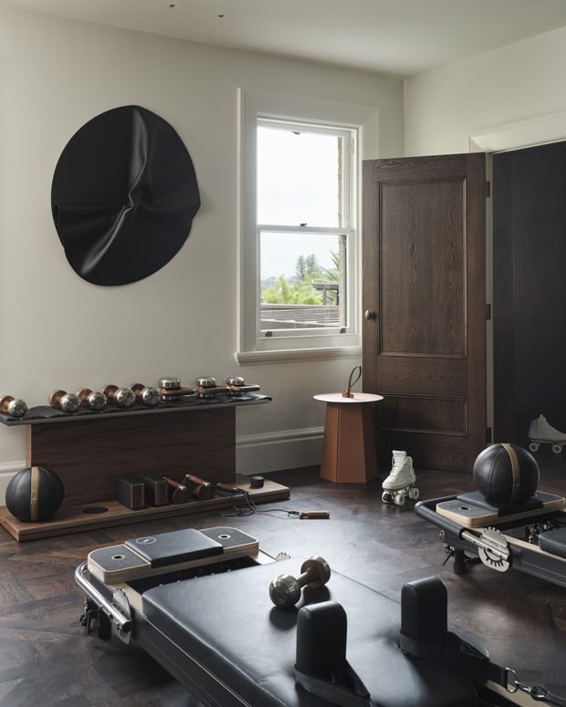 A refined home gym occupies a bright, traditionally detailed room with pale walls, dark timber floors, and a tall timber-panelled door. The space contains black leather Pilates reformer equipment, weights, exercise accessories, and a dark built-in storage console. A large sculptural black wall artwork and a small central side table add visual interest, while a sash window provides natural light and views of greenery outside.