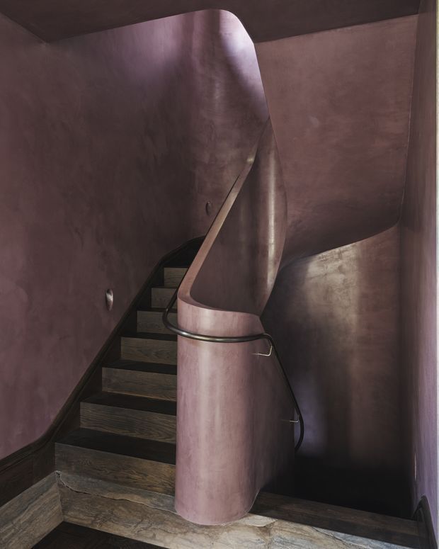 A sculptural enclosed stairway with dark timber treads and risers, surrounded by curved plaster or rendered walls in a muted mauve-brown finish. A sweeping central balustrade and rounded newel form create a dramatic, continuous architectural feature, with a dark metal handrail following the curve. The space is dimly lit and highly tactile, with an organic, monolithic character.