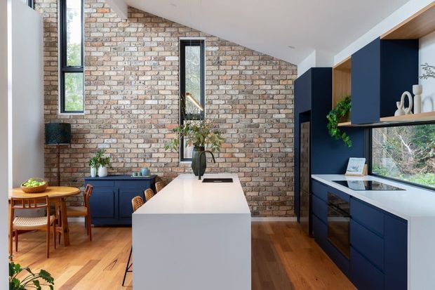 A bright contemporary kitchen with a central white island, deep navy cabinetry, timber open shelving, white benchtops, and a full-height exposed brick feature wall. The kitchen includes integrated appliances, a sink beneath a wide window, timber flooring, and a small adjoining dining area with a round timber table and woven chairs. A sloping ceiling and tall narrow windows add architectural interest.