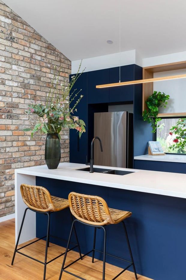 A contemporary kitchen interior featuring a deep navy island and cabinetry, white engineered-looking benchtop, stainless-steel refrigerator, black sink and tapware, and two woven rattan bar stools. An exposed warm brick wall contrasts with timber flooring and open timber shelving. A linear pendant light, indoor greenery, and a window add warmth and natural light.