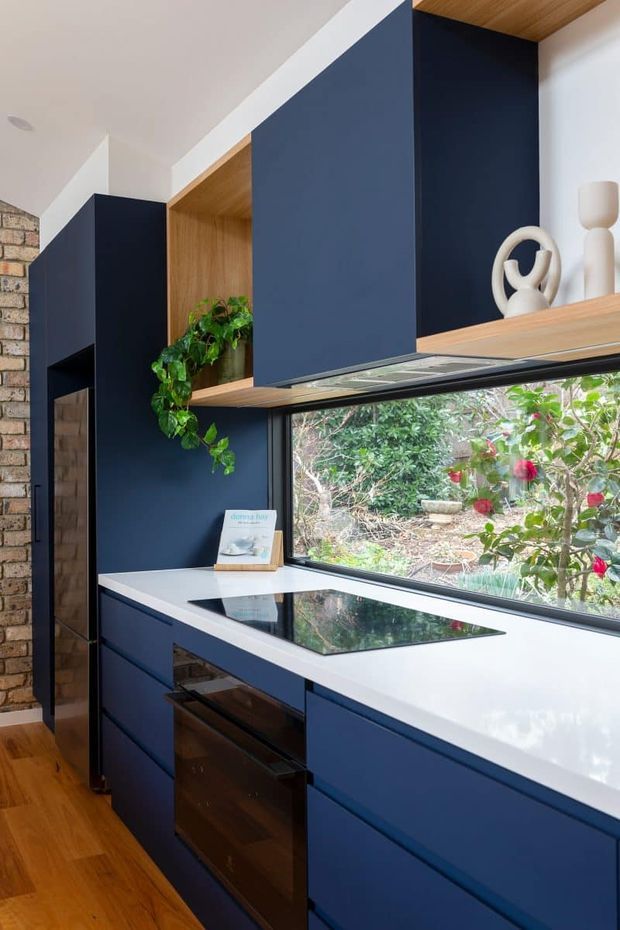 A narrow, contemporary kitchen with deep navy-blue built-in cabinetry, a white benchtop, and a black glass induction cooktop beneath a large horizontal window. Warm timber shelving, ceiling lining, and hardwood flooring contrast with the dark joinery, while exposed brick is visible at the left. Open shelving displays neutral ceramic objects and a trailing plant, with a leafy garden seen through the window.