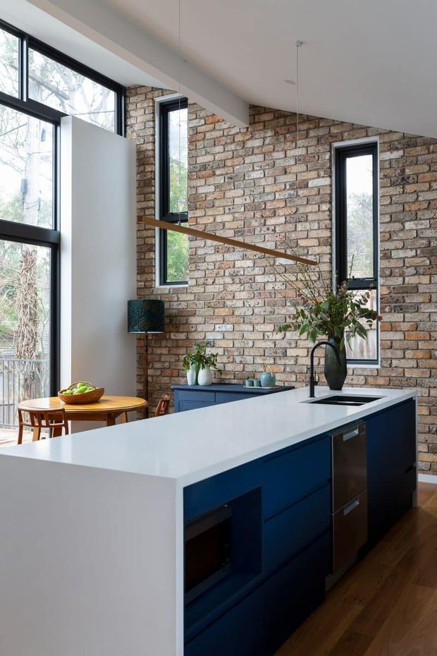 A bright open-plan kitchen and dining area with a large white island, deep navy-blue cabinetry, integrated appliances and sink, warm timber flooring, and an exposed recycled-brick feature wall. Tall black-framed windows bring in abundant natural light, while a small timber dining table and pendant lighting complete the space.