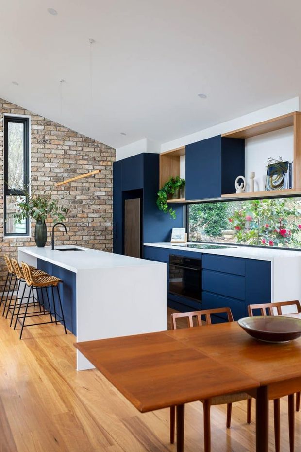 A bright open-plan kitchen and dining area with deep navy-blue cabinetry, a large white island with a black tap and integrated sink, warm timber flooring, and a matching timber dining table. An exposed light brick feature wall, timber open shelving, a long horizontal garden-facing window, and indoor plants add texture and warmth. Woven bar stools and mid-century-inspired dining furniture complete the space.