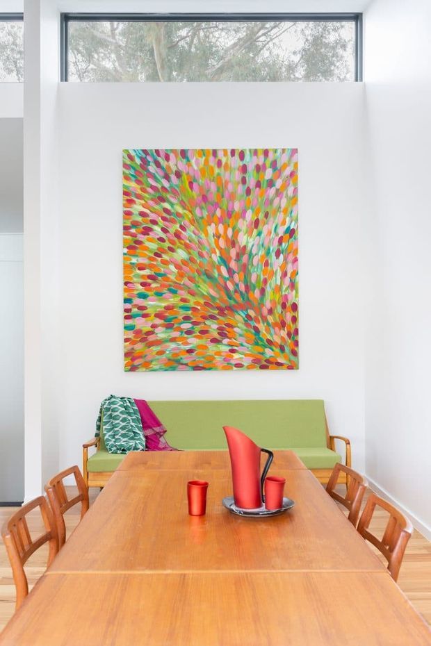 Bright dining room with a long warm timber dining table and matching wooden chairs, facing a green upholstered bench seat. A colourful abstract artwork is mounted on the white wall beneath a high horizontal window, creating a vibrant mid-century-inspired interior.