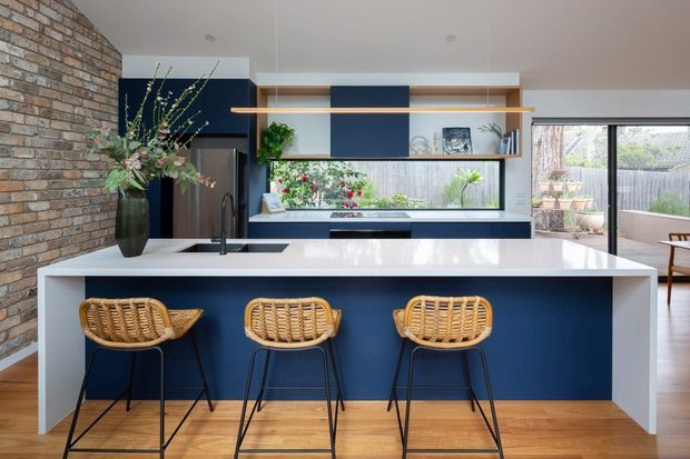 Bright contemporary kitchen with a large white island and deep navy cabinetry, featuring three woven rattan bar stools. A textured exposed brick wall contrasts with the timber flooring and clean white surfaces. Open shelving, a long horizontal window, pendant lighting, and a glimpse of an outdoor area complete the space.