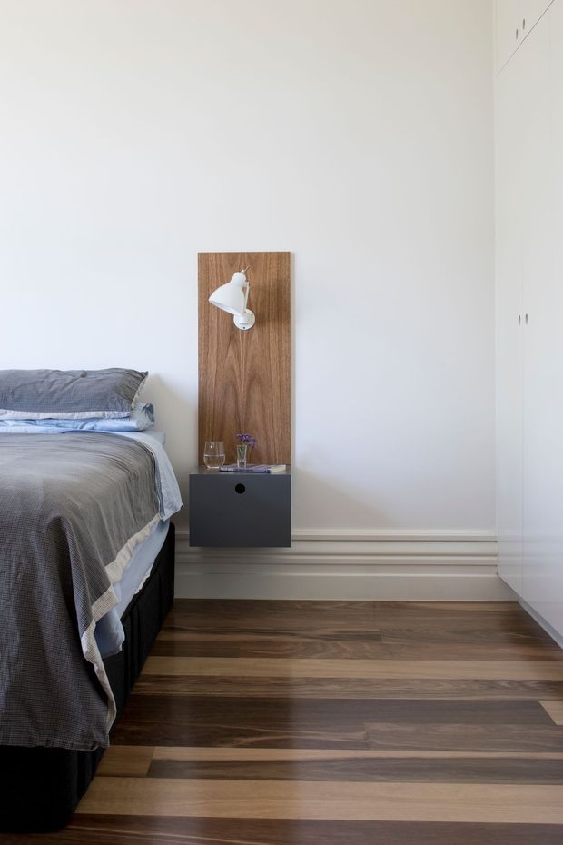 A calm minimalist bedroom with a partially visible bed dressed in grey bedding, a floating dark bedside drawer, and a vertical timber wall panel with a white reading lamp. White built-in wardrobes occupy the right side, while wide-plank timber flooring and simple decorative wall mouldings add warmth and detail.