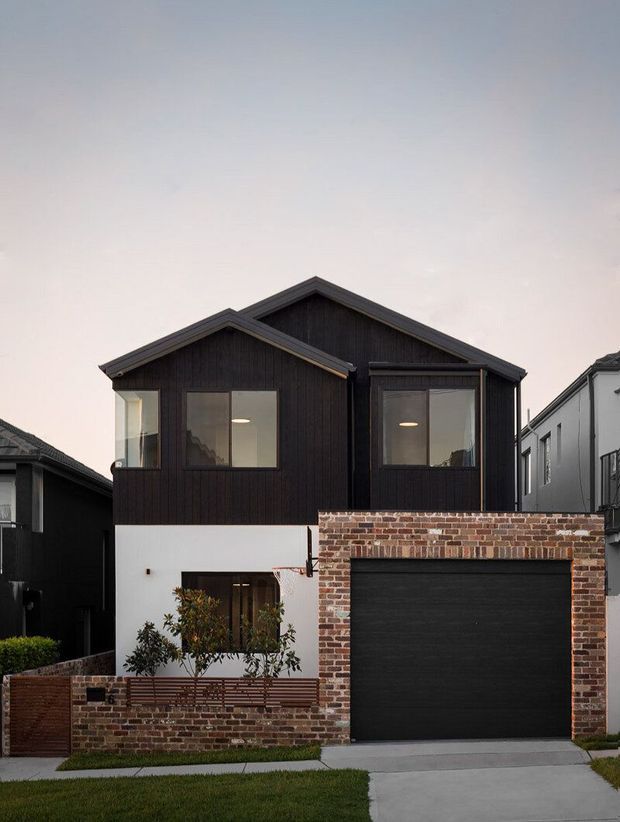 Street-facing exterior of a contemporary multi-level townhouse with a dark vertical-clad upper facade, white rendered lower level, prominent pitched gable roof forms, large windows, and a red-brown exposed brick garage and boundary wall. The facade includes a wide dark sectional garage door, small landscaped frontage, and concrete driveway.