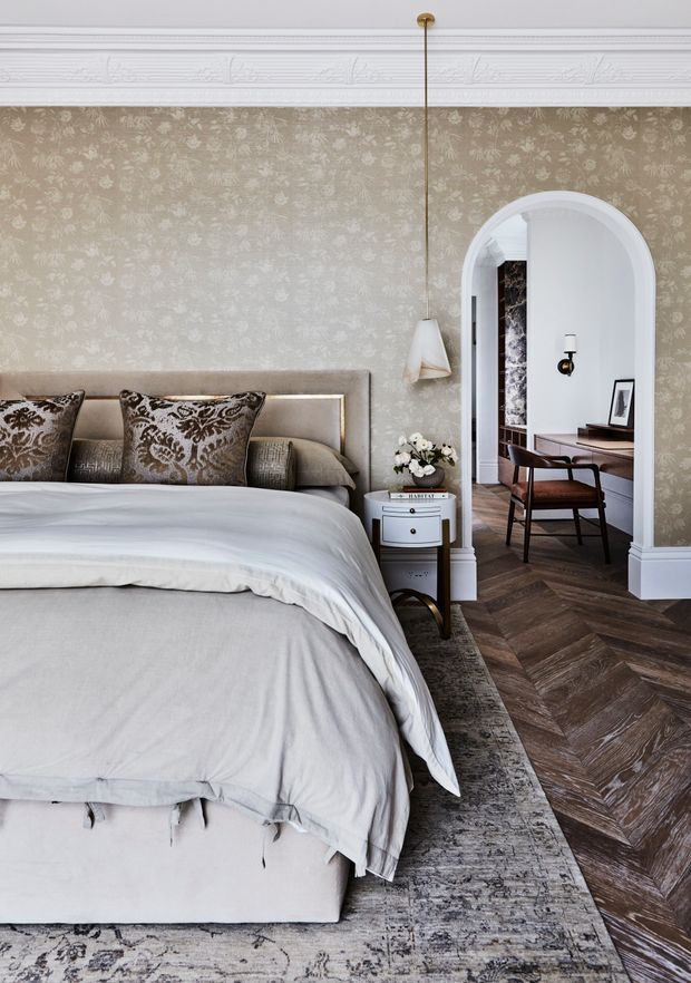 Elegant bedroom with a large upholstered bed, soft grey bedding, patterned accent pillows, floral wallpaper, ornate white ceiling moulding, and a herringbone timber floor. An arched opening leads to a compact adjoining workspace or sitting area, with a pendant light, bedside table, patterned rug, and refined traditional detailing.
