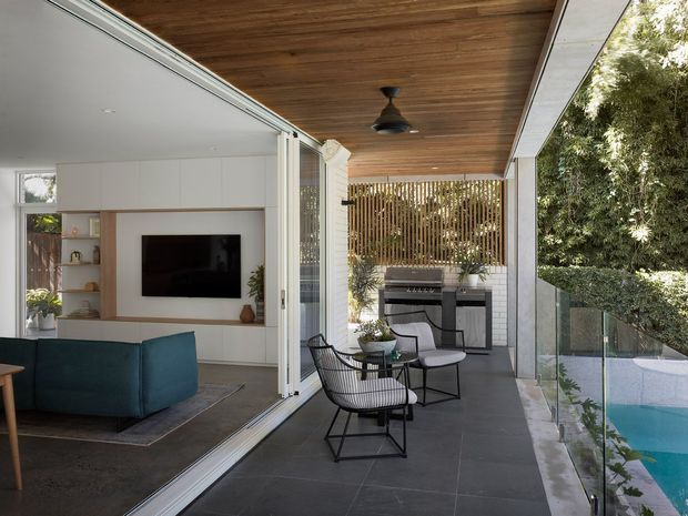 A covered outdoor terrace beside a swimming pool, connected to a contemporary living room through fully open glazed doors. The terrace has dark tiled flooring, a warm timber-lined ceiling with a ceiling fan, a glass balustrade, and black wire-frame lounge chairs around a small table. At the far end is a built-in outdoor kitchen or barbecue zone with a vertical timber privacy screen and greenery beyond. The living room visible inside includes white built-in cabinetry, open shelving, and a wall-mounted television.