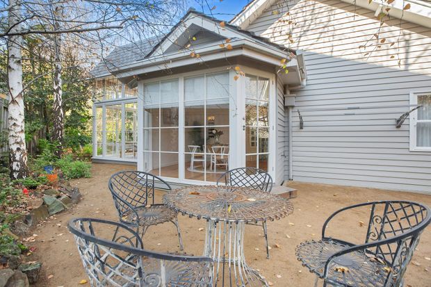 A bright exterior view of a cottage-style home with pale grey horizontal weatherboard cladding, a glazed sunroom or conservatory, and a small paved outdoor sitting area furnished with an ornate metal dining set. Mature birch trees, garden planting, and a pitched gabled roof contribute to a traditional, garden-oriented character.
