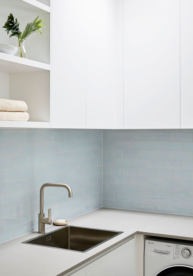 Bright, compact contemporary kitchen or kitchenette with white flat-panel cabinetry, open shelving, pale blue subway-tile splashback, a white benchtop, stainless-steel undermount sink and curved tap. A front-loading washing machine is integrated beneath the counter, with folded towels and a vase on the shelving.