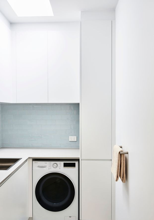 Bright, compact white laundry room with floor-to-ceiling flat-panel cabinetry, a concealed front-loading washing machine, pale blue-grey subway tile splashback, white benchtop, and an integrated sink at left. A skylight provides strong natural illumination, while a towel hangs from a wall-mounted rail on the tall cabinet.