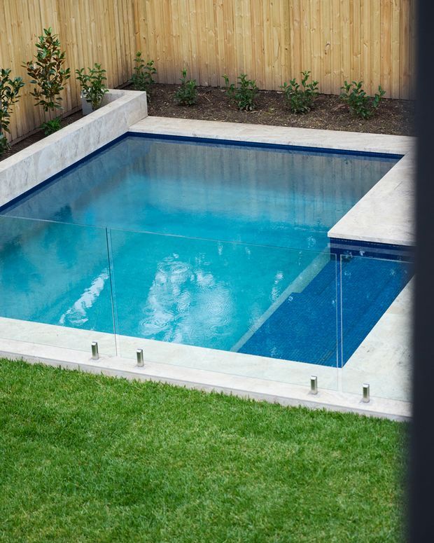 A compact rectangular swimming pool in a landscaped backyard, bordered by pale stone or concrete coping and clear frameless glass safety fencing. The pool has bright blue water with a darker blue tiled section, while timber fencing and small planted beds line the rear boundary. A neatly maintained lawn occupies the foreground.