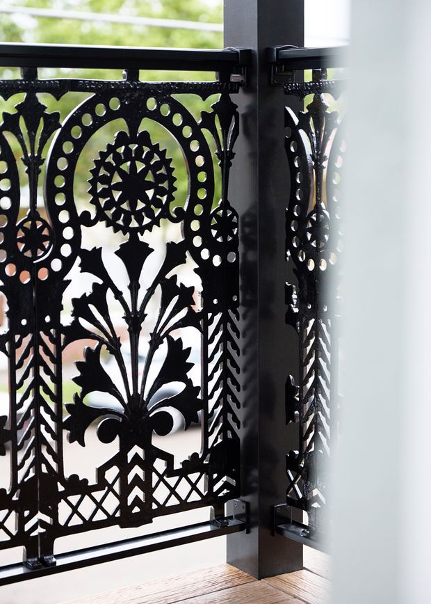 Close-up architectural detail of an ornate black metal decorative balustrade or screen, featuring intricate floral, botanical and geometric cut-out patterns. The panel is attached to a dark structural post with visible metal brackets and overlooks a bright exterior or void. Timber flooring is visible at the base, with strong contrast between the black metalwork and daylight beyond.