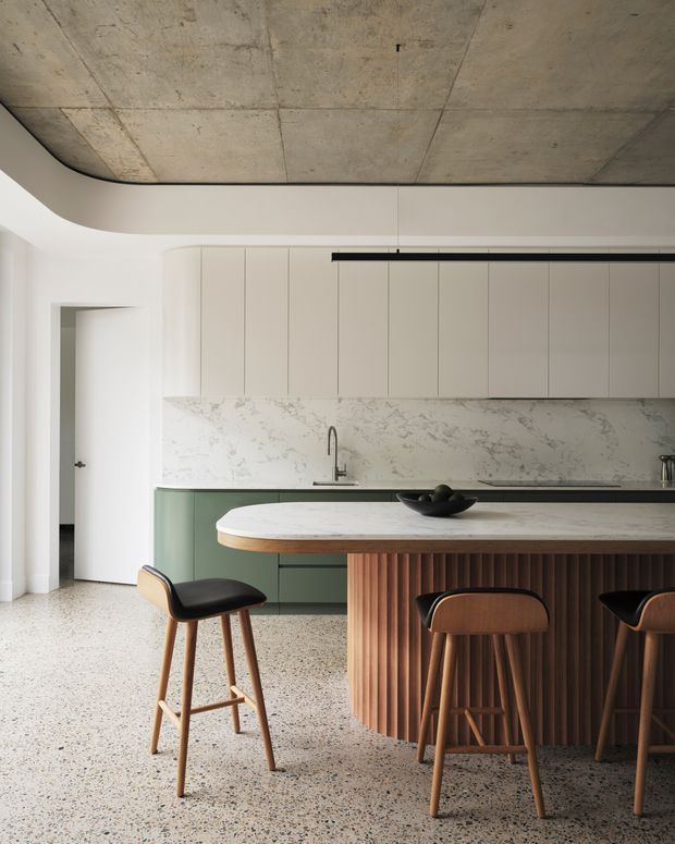 A refined contemporary kitchen with a large island featuring a white stone benchtop, curved timber edge, and vertically fluted timber-clad base. The kitchen includes muted green lower cabinetry, tall off-white cabinetry, a veined stone splashback, terrazzo flooring, and an exposed concrete ceiling. Minimal black linear lighting and timber-and-black bar stools complete the warm, modern interior.