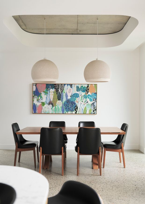 A bright, refined dining room with a long timber dining table surrounded by black leather-and-timber chairs. Two large spherical pendant lights hang above the table, with a colourful framed artwork on the white wall behind. The room features a polished exposed concrete ceiling recess and a terrazzo-style floor, creating a contemporary interior with mid-century influences.