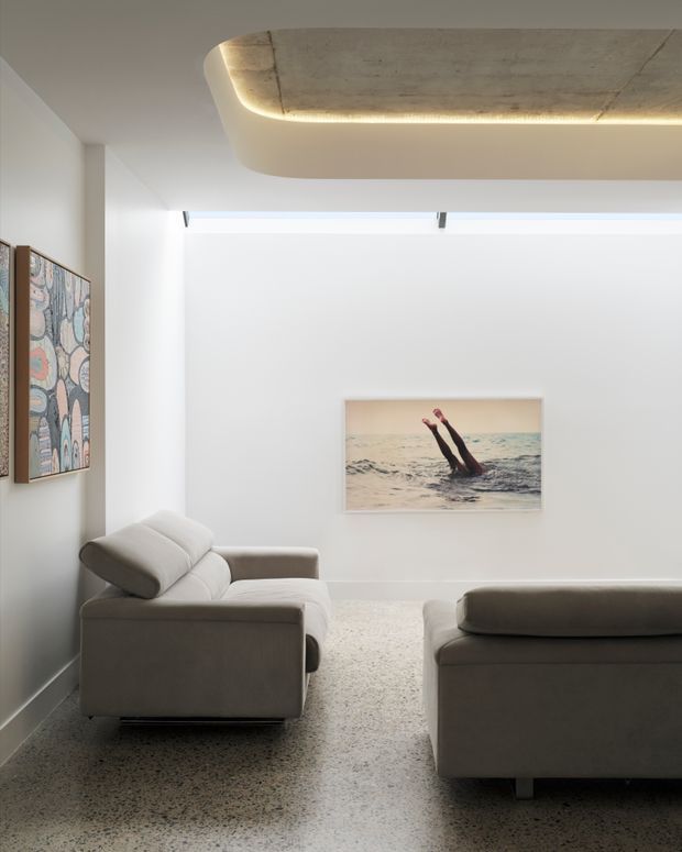Bright contemporary living room with two low-profile upholstered sofas, white walls, a polished terrazzo floor, and a sculptural recessed ceiling feature with concealed warm lighting. Abstract artwork and a photographic artwork add restrained visual interest.