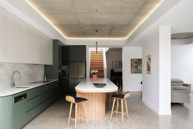 Contemporary open-plan kitchen with a central rounded island, dark green lower cabinetry, white upper cabinets, pale stone or terrazzo-style surfaces, and a marble-look splashback. Timber bar stools surround the island. A concrete-look recessed ceiling with concealed perimeter lighting adds a refined industrial touch, while the dining area, staircase and living room are visible beyond.
