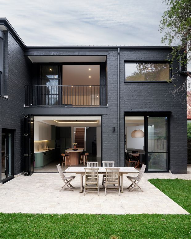 A contemporary two-storey courtyard-facing residence with a dark painted brick facade, flat roof, large black-framed windows and doors, and an upper-level balcony. The ground floor opens onto a pale stone-paved patio with an outdoor dining table and chairs, while the interior kitchen and dining spaces are visible through the openings. A small lawn borders the patio.