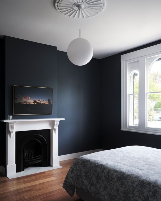A moody bedroom with deep charcoal walls, a white decorative fireplace mantel and black fire surround, polished timber flooring, a large bed with a dark patterned coverlet, and a white-framed arched window. A spherical pendant hangs beneath an ornate ceiling medallion, while framed artwork rests above the mantel.