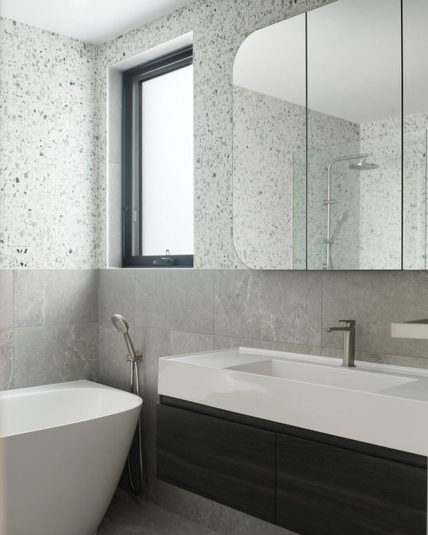 Contemporary bathroom featuring a freestanding white bathtub, wall-mounted dark timber vanity with integrated white basin, brushed metal tapware, large grey stone-look wall tiles, and terrazzo-style upper wall surfaces. A mirrored cabinet reflects the glass shower enclosure.