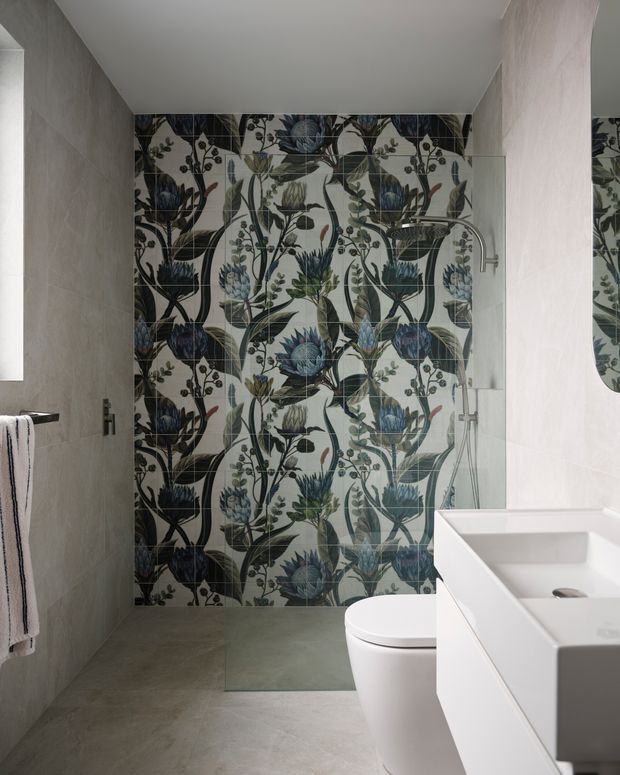 Contemporary bathroom featuring a full-height decorative botanical tile feature wall in dark blue, green, cream and black, with large-format grey wall and floor tiles. A frameless glass shower screen, wall-mounted shower fittings, white toilet and streamlined white vanity complete the space.