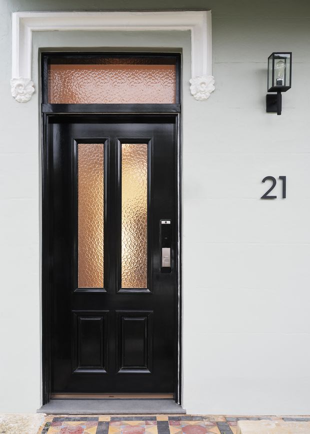 Close-up exterior view of a traditional black entrance door with raised panels, textured amber glass inserts, a contemporary smart lock, white decorative trim, wall-mounted lantern, house number 21, and a colourful patterned tiled threshold.