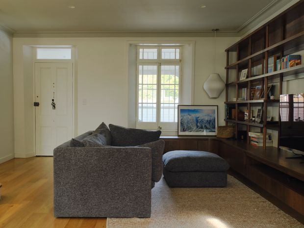 A refined living room with warm timber flooring, a charcoal upholstered sofa and ottoman, and extensive dark timber built-in shelving and cabinetry along one wall. The room features traditional panelled detailing, a white entry door, a large divided-light window, integrated display storage, artwork, and soft neutral finishes.
