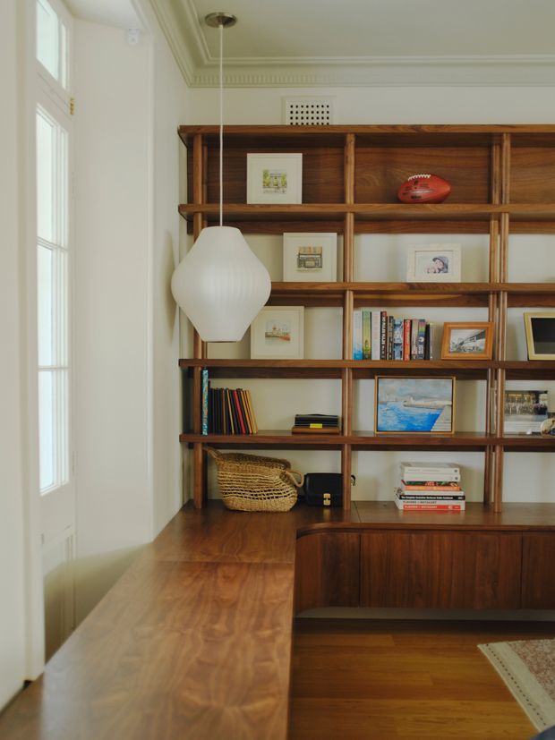 A warm, refined home office or study with extensive built-in dark timber shelving and cabinetry, displaying books, framed artwork, and decorative objects. A timber desk surface wraps around the shelving, with a sculptural white pendant light, white walls, detailed ceiling cornices, a glazed timber-framed door, and timber flooring.