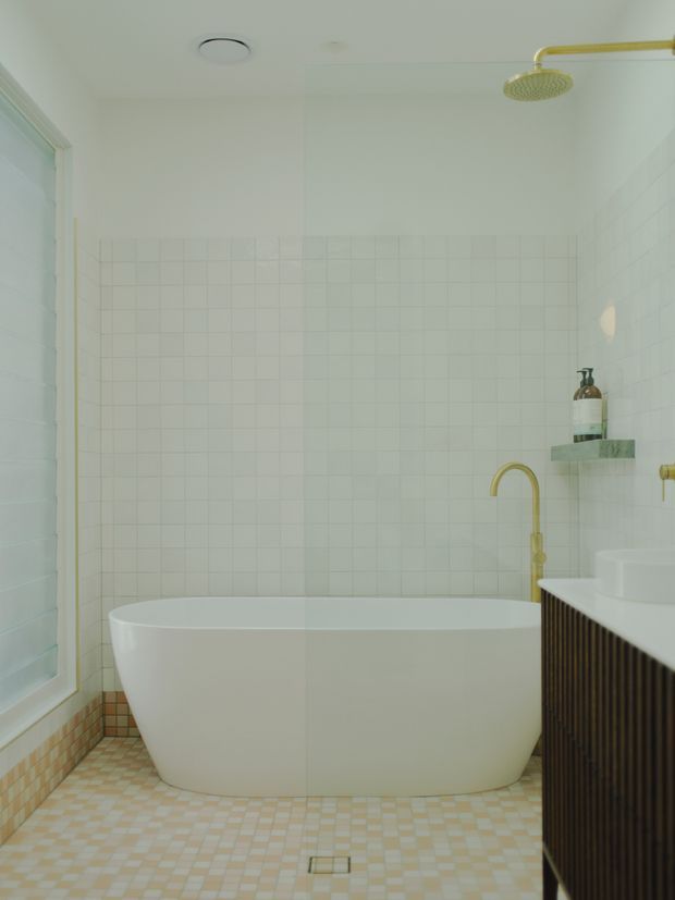 Bright, softly styled contemporary bathroom featuring a freestanding white bathtub, white square wall tiles, small checkered floor tiles, a white vanity with dark vertical fluted cabinetry, and brushed brass shower and tapware. A narrow window and recessed ceiling vent add natural light and ventilation.