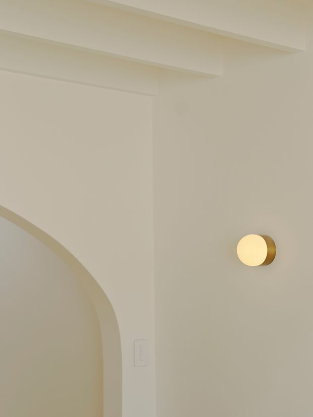 Close-up of a softly lit interior wall in warm off-white plaster, featuring a curved arched opening at left, layered ceiling coving or recessed architectural detailing above, and a small round wall sconce with a warm glowing light. The image is minimalist and focused on architectural surfaces and lighting.
