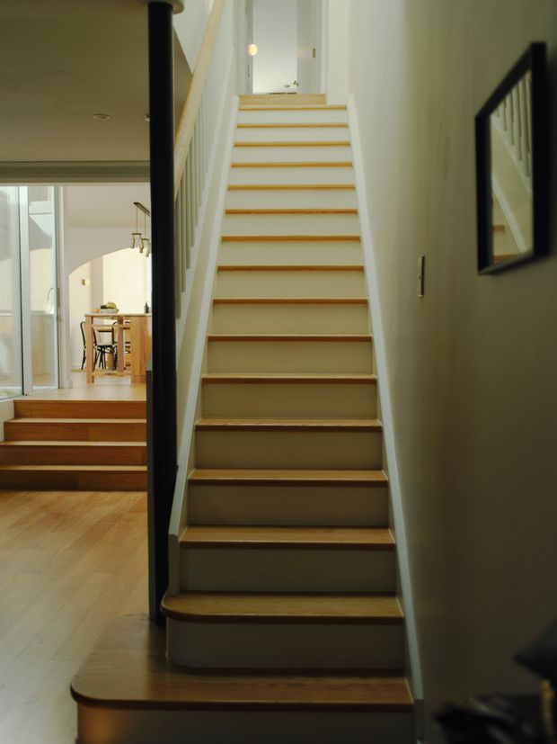 Interior view focused on a narrow staircase with warm timber treads, painted light walls, and white balustrade spindles. A black vertical handrail frames the stair, while an adjacent open-plan dining area and timber flooring are visible to the left. A wall mirror reflects part of the stairway.