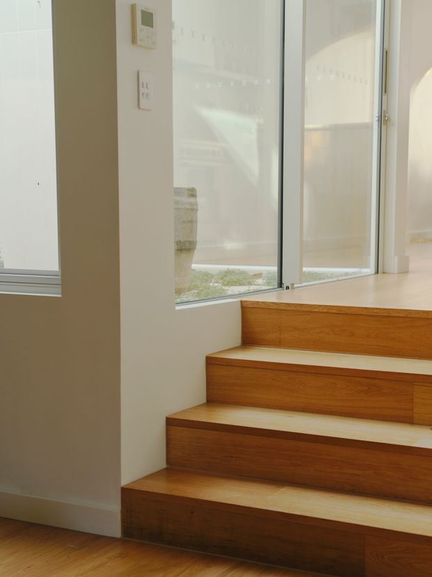 Bright minimalist interior showing a short flight of built-in timber stairs with open risers and broad wooden treads, beside large floor-to-ceiling glazed windows. White painted walls, slim light-coloured window frames, and warm natural timber create a clean, contemporary stairway setting.