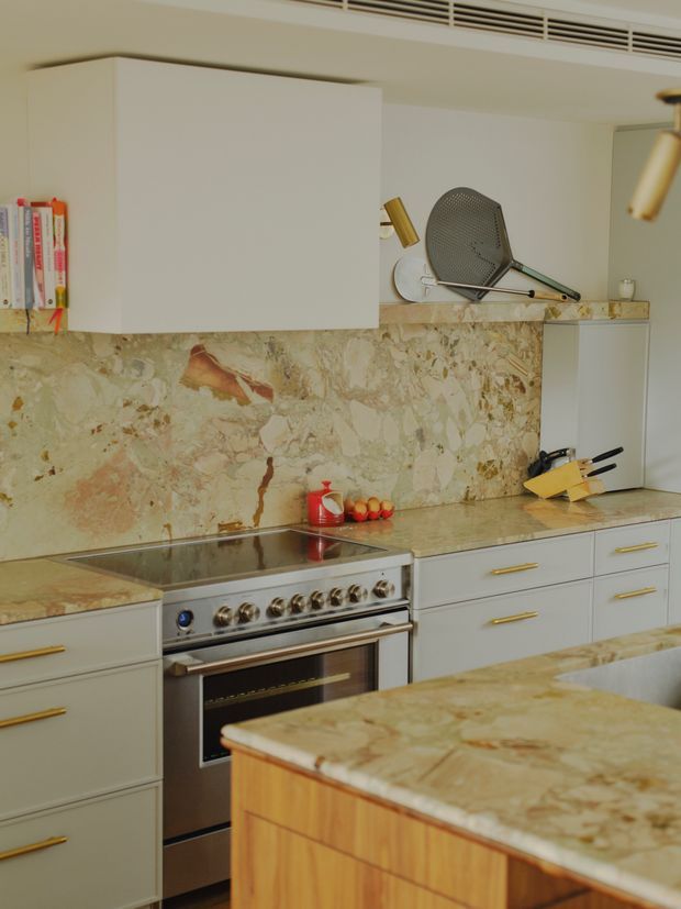 A warm, compact kitchen with pale grey-white flat-panel cabinetry, brass pulls, a large patterned natural-stone backsplash and matching stone benchtops. The kitchen includes a freestanding stainless-steel range, induction cooktop, open shelving with books and decorative objects, and a timber-sided island in the foreground.