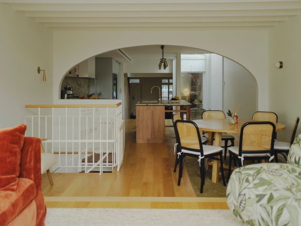 Open-plan residential interior viewed from a living area toward a dining space and kitchen. The room features a broad rounded arch opening, white walls and ceiling beams, warm timber flooring, a timber kitchen island, a round dining table with cane-backed black-framed chairs, and a white stair balustrade. Colourful upholstered seating and leafy-patterned furniture add a playful accent.