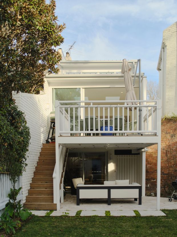 Rear exterior view of a compact multi-level home extension with a raised timber deck and white balustrade. The upper level has glazed doors, outdoor dining furniture and a closed umbrella, while the lower level forms a covered outdoor lounge or entertaining area. White-painted brick walls, timber stairs, exposed brick, paved flooring and a small lawned garden surround the structure.