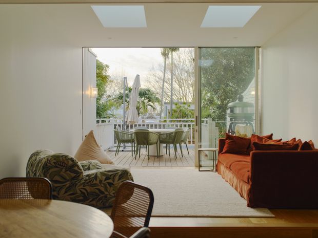 Bright open-plan living and dining area with a warm, relaxed interior. A large upholstered orange sofa and patterned lounge chair face the room, while a round timber dining table and woven chairs sit in the foreground. Full-height glazed doors open onto a furnished outdoor terrace with a dining setting, umbrella, and lush planting. The room has pale walls, recessed ceiling lights, a light carpet, and timber flooring near the terrace threshold.