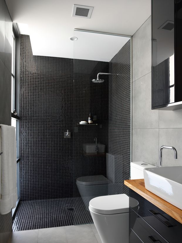 A compact contemporary bathroom featuring a walk-in shower and toilet enclosed in dark small-format mosaic tiles. Large light grey wall tiles, a white vessel basin on a timber vanity benchtop, black cabinetry, chrome tapware, a wall-mounted mirror, and recessed ceiling ventilation create a high-contrast, modern finish.