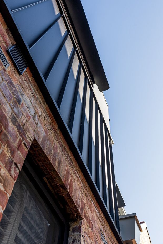 Upward-looking close-up of a contemporary building facade, showing weathered red brick masonry with arched openings, dark blue-grey metal-framed glazing and projecting roof or sunshade elements against a clear sky.