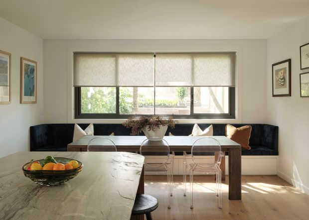 Bright, refined open-plan dining and living area with a large stone-topped dining table in the foreground, transparent acrylic dining chairs, and a built-in dark upholstered banquette beneath a wide window. Light timber flooring, pale walls, framed artwork, soft roller blinds, and a simple central pendant create a calm contemporary setting.