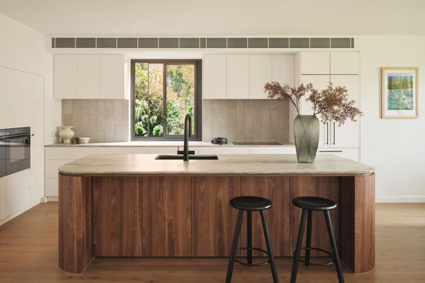 Bright contemporary kitchen featuring a large curved island with dark timber cabinetry, a pale stone benchtop, integrated appliances, white flat-panel cabinetry, a textured stone backsplash, and a black gooseneck tap. Two black stools sit at the island, while a central window brings in garden views and a slim horizontal ventilation grille runs above the wall cabinets.