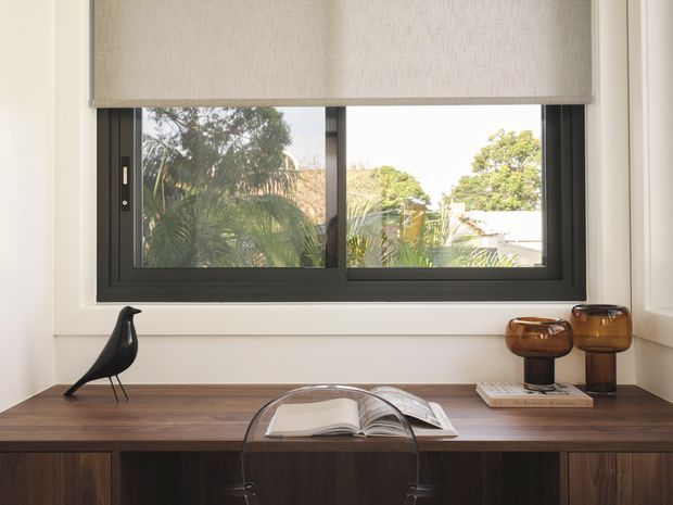 A calm, refined interior vignette featuring a dark timber built-in console beneath a large black-framed window. A roller blind covers the upper portion of the glazing, with tropical greenery visible outside. Decorative amber glass vessels, an open book, and a black bird sculpture sit on the console, while a transparent chair is positioned in front.