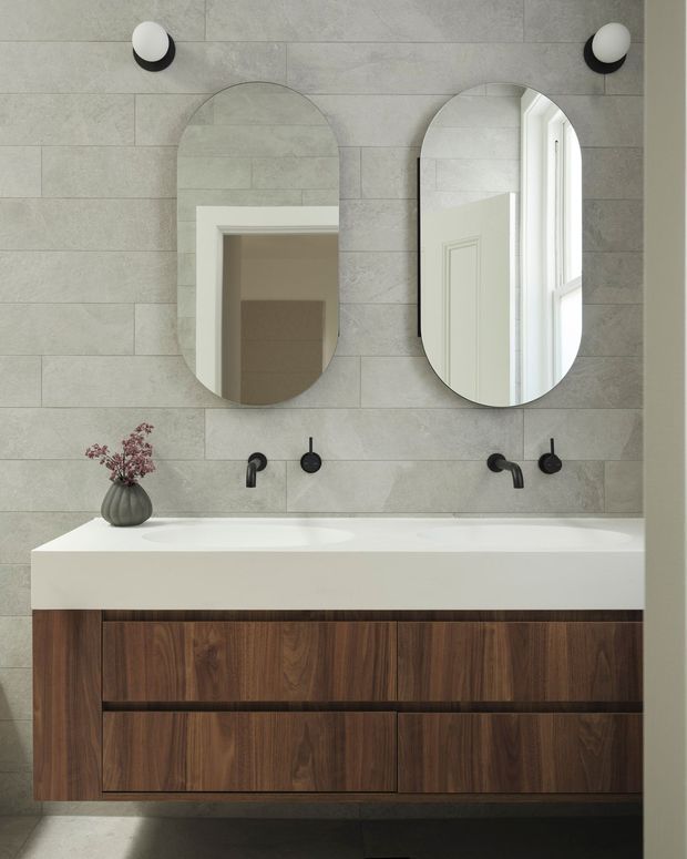 Contemporary bathroom vanity with a wide white integrated benchtop and basin, dark timber-grain cabinetry, pale grey large-format tiled walls, matte black wall-mounted tapware, and two rounded-arch mirrors. A small vase provides a subtle decorative accent, while a doorway and window are reflected in the mirrors.