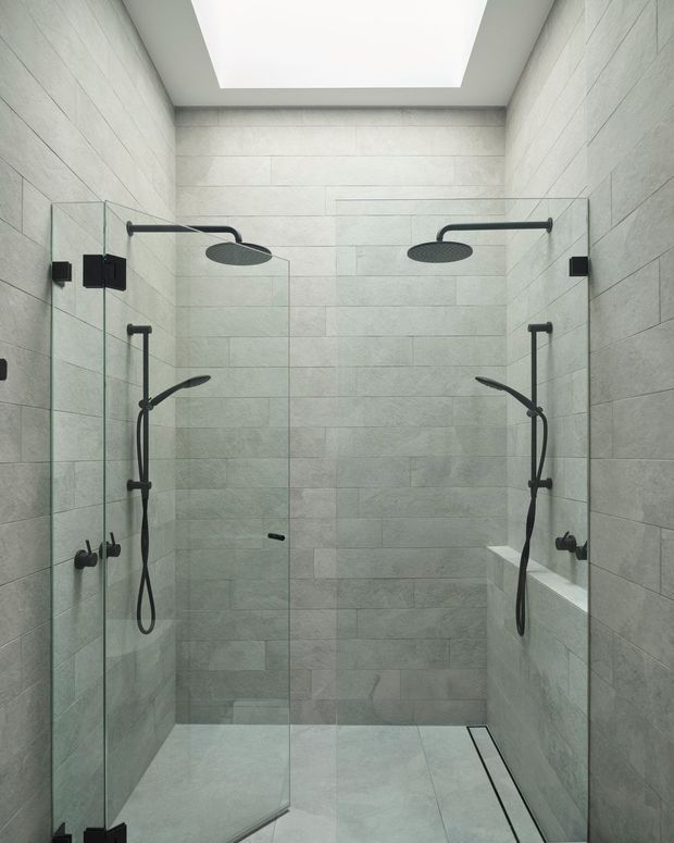 A contemporary walk-in double shower enclosed by clear frameless glass panels. The shower walls and floor are clad in large-format pale grey stone-look tiles, with matte black dual overhead rain showers, handheld fittings, controls and hardware. A narrow linear drain runs along the floor, and a large ceiling skylight or lightwell provides bright diffuse illumination.