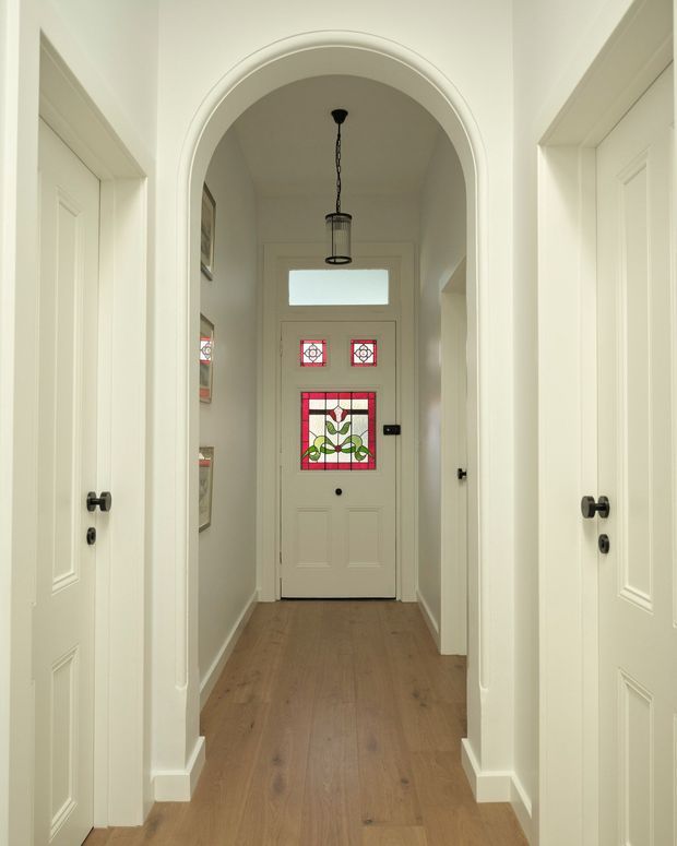 A narrow, bright residential hallway leading to a front door, framed by a prominent white arched opening. White paneled internal doors with dark round hardware line both sides, while the timber floor adds warmth. The entrance door features decorative stained-glass panels and a small transom window, with a hanging lantern-style light fitting centered beneath the arch.